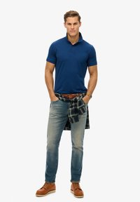 Polo navy con colletto, jeans azzurri, e scarpe in pelle marroni. Una camicia a quadri annodata in vita aggiunge un dettaglio a strati.