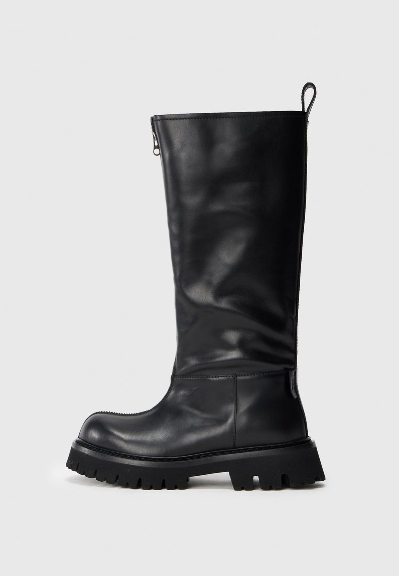 Botte noire en cuir haute avec semelle épaisse à crampons, bout arrondi et une languette à l'arrière, contre un fond clair et uni.