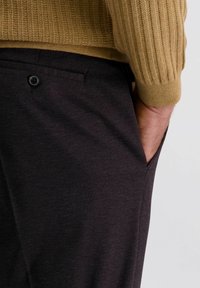 Mann in strukturierten dunklen Hosen mit einer Gesäßtasche mit Knopf, Hand in der vorderen Tasche, und einem sichtbaren Ärmel eines beigen RippPullovers.