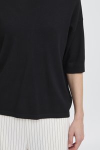 Maglione nero oversize lavorato a maglia con maniche corte. Texture liscia, vestibilità rilassata. Abbinato a pantaloni bianchi a righe verticali.