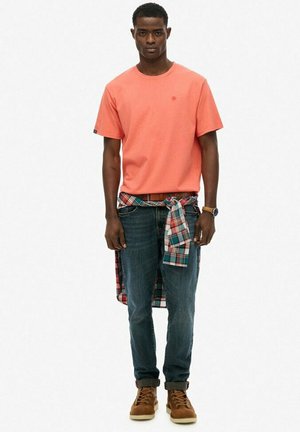 Koralfarvet t-shirt med korte ærmer, blå jeans og brune støvler. En ternet skjorte bundet om taljen tilføjer et lagdelt detalje.