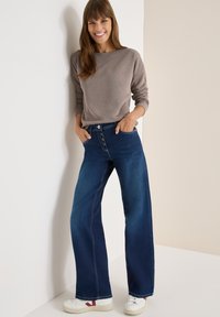 Graues, geripptes Langarmoberteil kombiniert mit hochtaillierten, weiten, dunkelblauen Jeans, die einen Knopfverschluss und kontrastierende Nähte aufweisen. Weiße Sneakers.