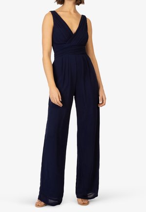 Combinaison bleu marine sans manches avec décolleté en V profond, corsage plissé, jambes larges et taille ajustée portée avec des sandales beige à bouts ouverts sur un mannequin.