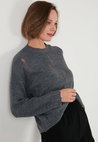 Grauer gestrickter Pullover mit strukturiertem Finish, der absichtlich distressed Details und einen Rundhalsausschnitt aufweist. Kombiniert mit dunklen Hose mit elastischem Bund.