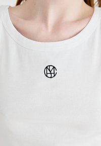 Biała, żebrowana koszulka bez rękawów z okrągłym dekoltem, zdobiona małym, czarnym haftowanym logo na środku. Miękka faktura materiału.