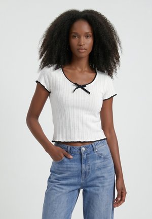 Femme aux cheveux bouclés portant un haut blanc côtelé à manches courtes avec bordure noire et un jean taille haute bleu clair, debout avec une main dans la poche.