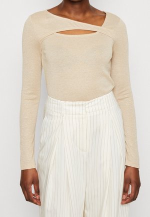Haut beige à manches longues avec un détail découpé au niveau du col, en tissu brillant, associé à un pantalon blanc à rayures.