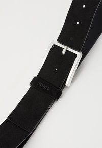 Zwarte suède riem met een zilveren metalen gesp, voorzien van meerdere gaten voor aanpassing en een logo op de band gestempeld.