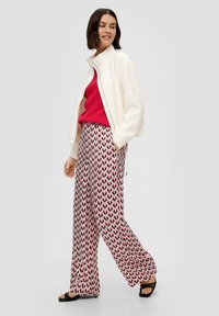 Weißer Reißverschluss-Bomberjacke, rosa gerippter Pullover, gemusterte weit geschnittene Hose mit pinkem und schwarzem Zickzackdesign und schwarzen Riemchensandalen.