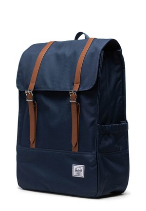 Mørkeblåt stof rygsæk med brune læderremme, sølvbeslag, klaplukning, sidelomme og et hvidt logomærke på den nederste front.