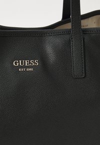 Czarna skórzana torba tote z teksturowaną powierzchnią, podwójnymi uchwytami oraz tłoczonym złotym logo "GUESS" z napisem "EST 1981" poniżej.