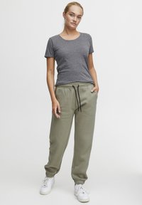 Graues Baumwoll-T-Shirt mit kurzen Ärmeln, kombiniert mit olivgrünen Jogginghosen mit elastischem Bund und schwarzem Kordelzug. Weiße Sneakers.