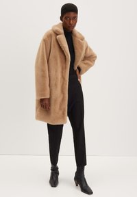 Cappotto in pelliccia sintetica color sabbia con colletto a punta, dotato di tasche laterali e una vestibilità rilassata. Indossato sopra un dolcevita nero e pantaloni neri aderenti.