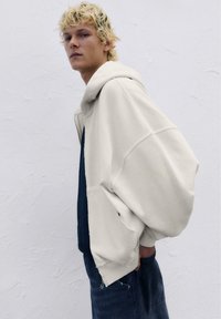 Übergroßer beiger Hoodie mit einer großen Kapuze, gerippten Bündchen und rohem Saum. Getragen über einem dunklen Hemd mit Knöpfen, kombiniert mit dunklen Jeans.