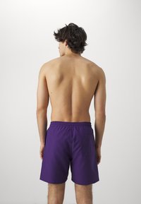 Bañador morado con cinturilla elástica y bolsillo trasero, visto desde atrás. El material parece ligero y suave. Piel visible en la espalda.
