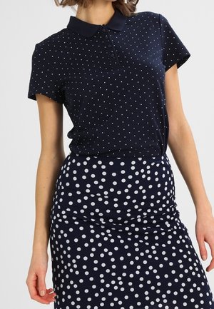 Mujer que lleva una camisa tipo polo azul marino con pequeños lunares blancos y una falda azul marino con grandes lunares blancos.