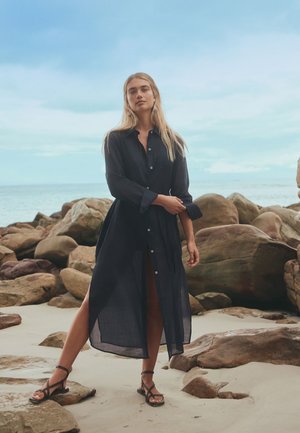Nő hosszú fekete gombos ruha és szandál visel, mezítláb áll homokos tengerparton nagy kövek között, részben felhős ég alatt.