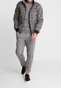 Man som går med grå rutmönstrad jacka, grå turtleneck-tröja, grå korta byxor och svarta sneakers med vita sulor.