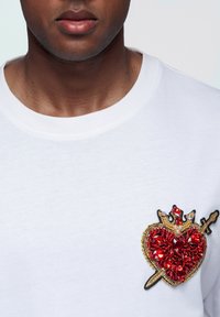 Weißes T-Shirt mit einem herzförmigen Edelsteinmuster aus roten Strasssteinen, goldenen und schwarzen Akzenten sowie einem Schwert, auf der linken Brustseite platziert.