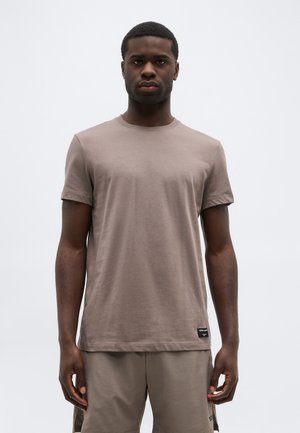 Νέος άνδρας με κοντά μαλλιά στέκεται προς τα εμπρός φορώντας ένα απλό ταπεζ ταμπά κοντομάνικο μπλουζάκι και αντίστοιχα σορτς, με φόντο λευκό.