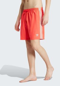 Koraļļu krāsas peldēšanas šorti ar baltām sānu svītrām un aukliņu jostu. Apakšējā kreisajā kājā redzams Adidas logotips. Viegls audums.