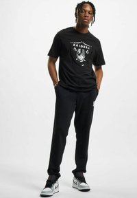 Svart bomulls T-skjorte med Raiders grafikk på fronten, kombinert med svarte joggebukser og hvite sneakers med svarte detaljer.