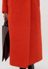 Lång orange ullkappa med texturerad finish, med en sidoficka, bärs tillsammans med en mörkbrun läderväska.