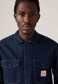 Chemise en denim boutonnée, bleu foncé avec deux poches poitrine, boutons noirs, coutures blanches et une étiquette rouge sur la poche gauche.