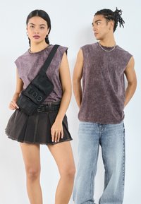 To personer har på seg avslappede antrekk; den kvinnelige modellen har en lilla, ermeless topp og et svart skjørt med frynser, mens hun holder en svart crossover-veske. Den mannlige modellen har på seg en matchende lilla ermeless topp og lyseblå jeans.