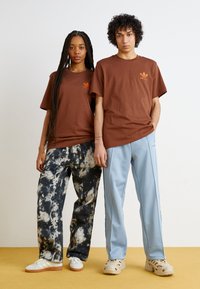 Bruna bomull t-shirts med en orange logotyp, tillsammans med blåa träningsbyxor och svart-vita tie-dye-byxor. Neutrala sneakers fulländar looken.