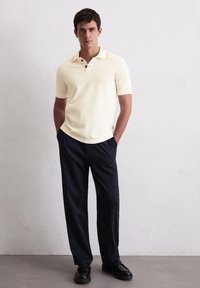 Koszulka polo z krótkim rękawem w jasnym żółtym kolorze, z klasycznym kołnierzykiem i trzema guzikami, zestawiona z ciemnoniebieskimi spodniami i czarnymi butami.
