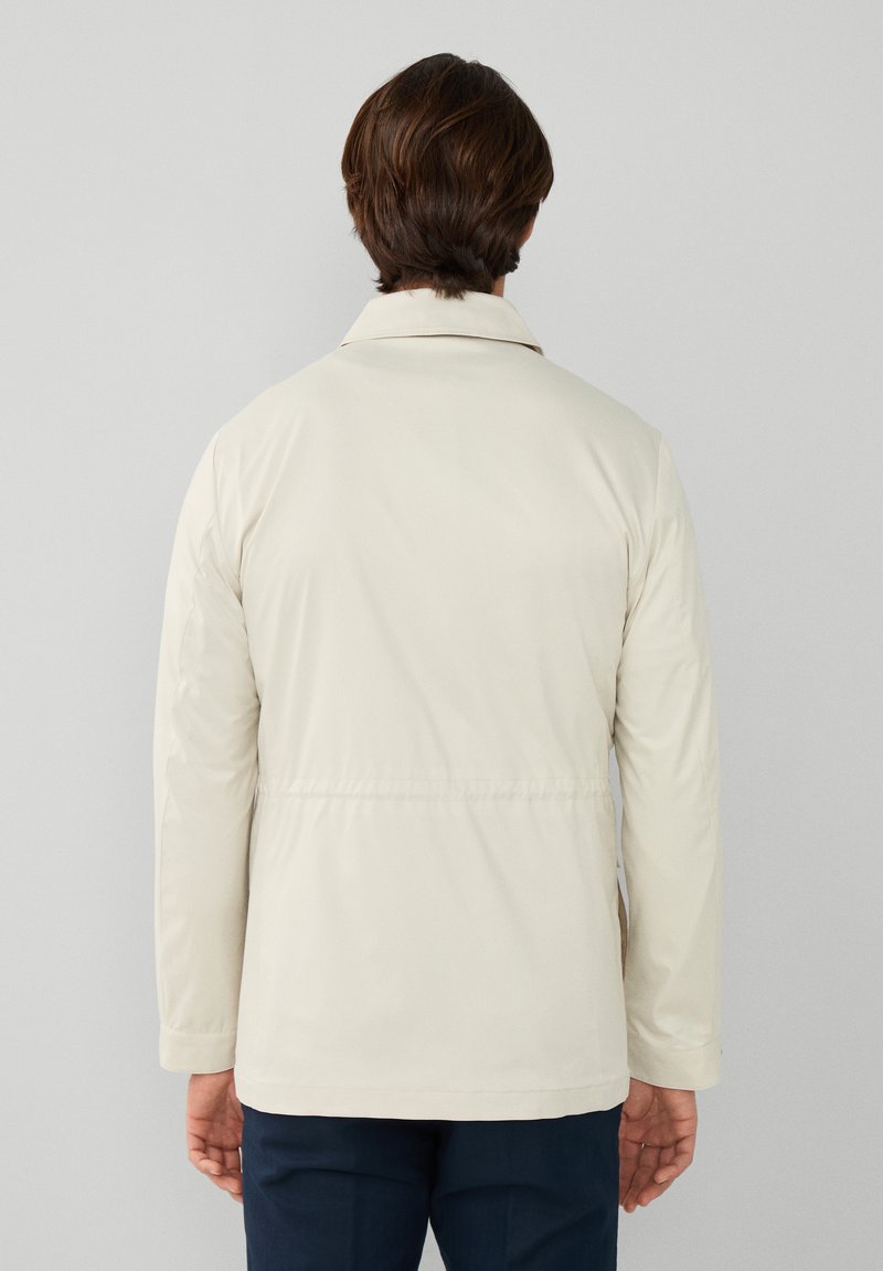 Veste beige clair au toucher doux, avec un col, des manches longues et une taille cintrée. La vue arrière montre un design minimaliste.