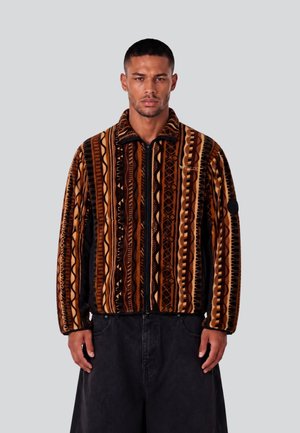 Mann trägt eine braun-schwarze gemusterte Reißverschlussjacke mit vertikalen Tribal-Designs und schwarze weite Hosen, steht vor einem schlichten grauen Hintergrund.