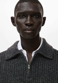 Ciemnoszary sweter dziergany na drutach z zamkiem błyskawicznym i wysokim kołnierzem, noszony na białej koszuli zapinanej na guziki, z teksturowanym prążkowaniem i metalowymi elementami.