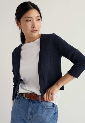 Femme portant un cardigan court bleu marine par-dessus un haut blanc avec un jean bleu et une ceinture marron, regardant vers sa gauche sur un fond uni.