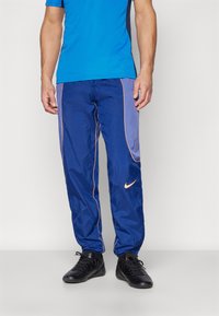 Blaue Jogginghose mit lila Akzenten, ausgestattet mit einem elastischen Bund, seitlichen Taschen und einem kleinen orangefarbenen Nike-Logo am Oberschenkel. Strukturierter Stoff.