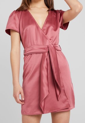 Mujer que lleva un vestido corto de satén en color rosa polvo, con mangas cortas y un amplio cinturón atado a la cintura.