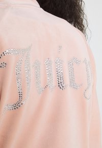 Rosa velour-hoodie med silverfärgade rhinestone-ordet "Juicy" på baksidan. Mjuk textur med en något figursydd design.