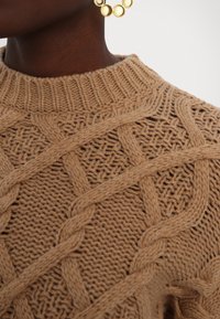 Personne à la peau foncée portant un pull torsadé beige avec un col côtelé et une boucle d’oreille ronde dorée imposante sur fond blanc.