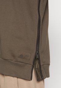 Gros plan sur un tissu marron avec une fermeture éclair latérale partiellement ouverte révélant la doublure intérieure, portant de petits logos Adidas et Parley près de l'ourlet.