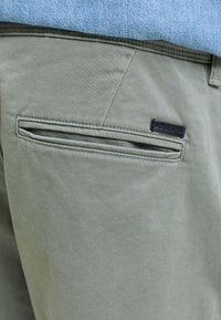 Rohelistest pükstest tagatasku lähedalt väikese musta Jack & Jonesi sildiga, seljas helesinine top.