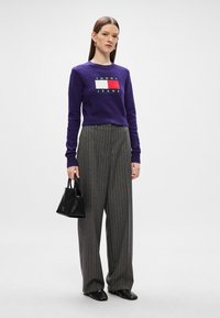 Μωβ κοντό πουλόβερ με το λογότυπο "TOMMY JEANS", γκρι ριγέ παντελόνι φαρδύς γραμμής και μία μικρή μαύρη τσάντα. Λείος υφασμάτινος σκελετός και μοντέρνα εφαρμογή.