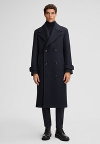 Marineblauer Wollpeacoat mit breiten Revers, doppelter Knopfleiste und großen schwarzen Knöpfen. Die Ärmel haben mit Knöpfen verzierte Akzente. Über einem schwarzen Rollkragenpullover getragen.