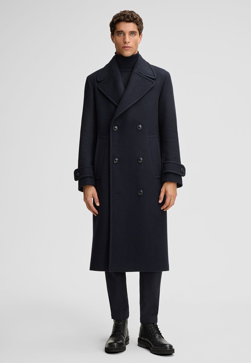 Marineblauer Wollpeacoat mit breiten Revers, doppelter Knopfleiste und großen schwarzen Knöpfen. Die Ärmel haben mit Knöpfen verzierte Akzente. Über einem schwarzen Rollkragenpullover getragen.