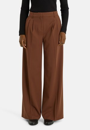 Pantalones marrones de pierna ancha con pliegues delanteros, hechos de tela suave. Combinados con una camiseta ajustada negra de tejido acanalado y calzado negro.