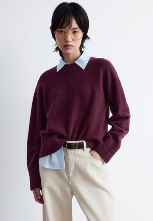 Burgunderfarbener Strickpullover mit Rundhalsausschnitt über einem hellblauen Hemd mit Kragen, kombiniert mit beigen weit geschnittenen Hosen und einem braunen Gürtel.