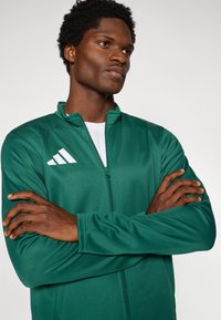 Πράσινο μπουφάν με φερμουάρ, κατασκευασμένο από λείο ύφασμα, που διαθέτει λευκή λεπτομέρεια με τρεις ρίγες στη λογότυπο στο αριστερό στήθος και σχέδιο με ψηλό γιακά.