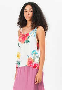 Blomstret tank top i hvid, med levende røde, lyserøde, gule og blå blomster. Glat stof, scoop-halsudskæring og løs pasform, parret med en lyserød nederdel.