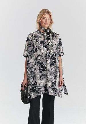 Mujer rubia lleva un top suelto con estampado floral en blanco y negro con un lazo en el cuello, pantalones negros y sostiene un bolso negro texturizado.