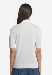 Vit kortärmad polo-tröja med mjuk textur, ribbad krage och en figursydd design. Visar en ren, minimalistisk bakvy.